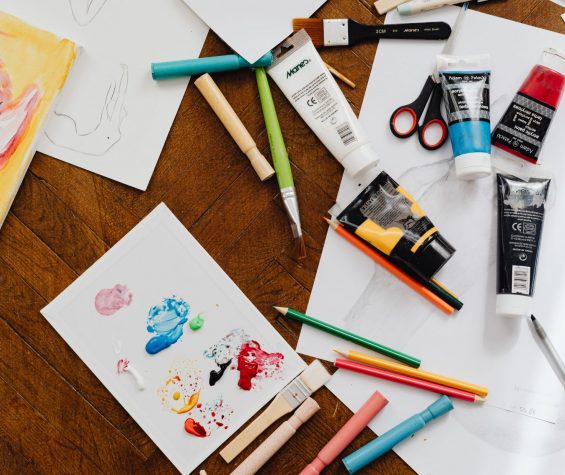 Collection of art materials including paints, pencils, paper and the start of art creations on white paper, spread across a dark wooden table.