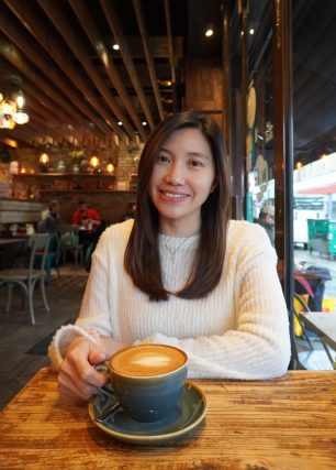 Gigi Chan, Art Psychotherpist from The Fountain Therapy Trust. Dark haired woman in white jumper seated at cafe table with coffee mug.